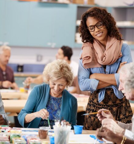 A cheerful group of Group Of Retired Seniors Attending Art Class In Community Centre With Teacher painting.